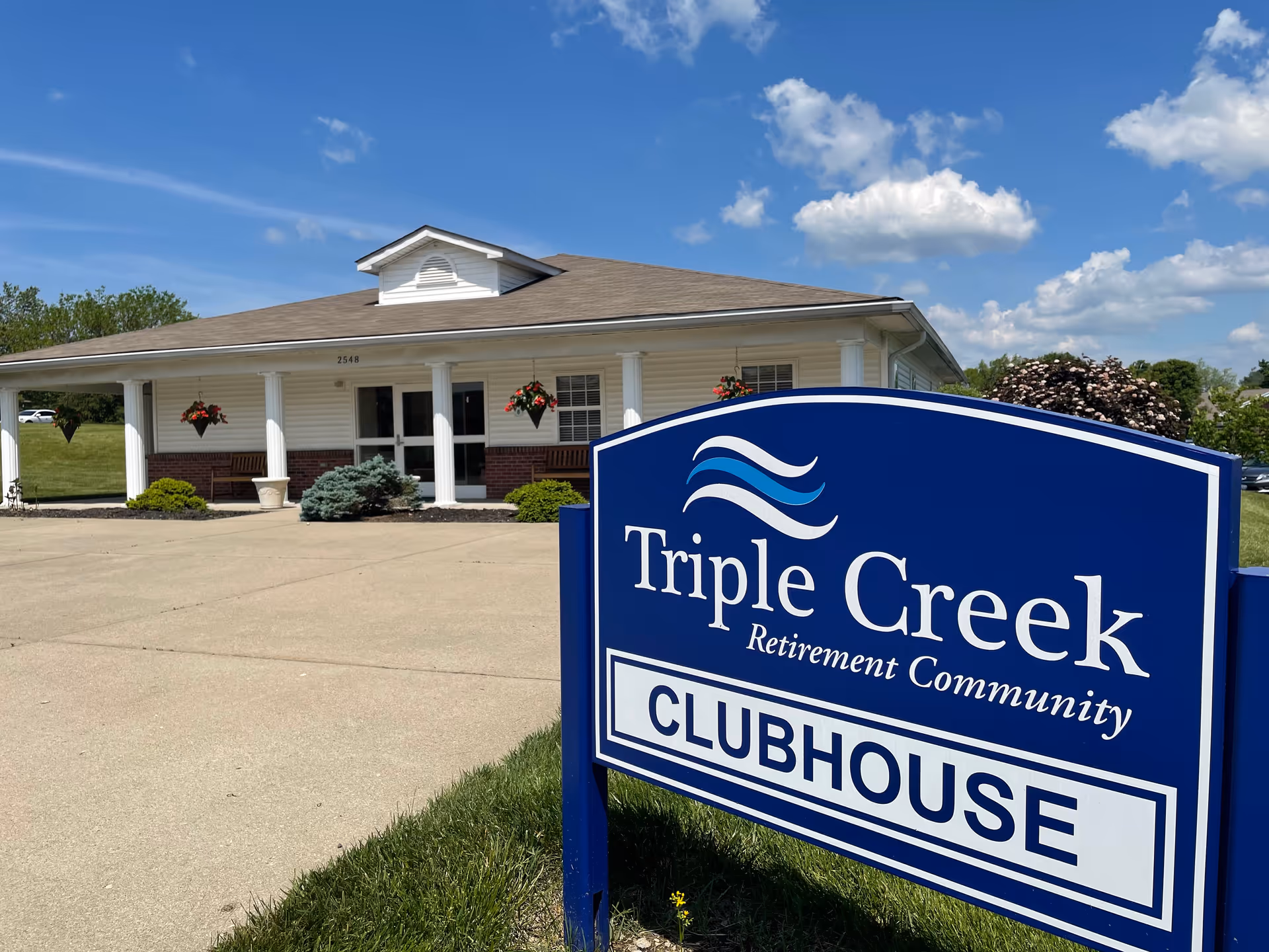 Exterior view of the Triple Creek Retirement Community clubhouse building on a sunny day with a clear blue sky and some clouds. The building has white siding, a brown roof, and white columns supporting a covered porch with hanging flower baskets. A large blue sign in the foreground reads 'Triple Creek Retirement Community CLUBHOUSE'.