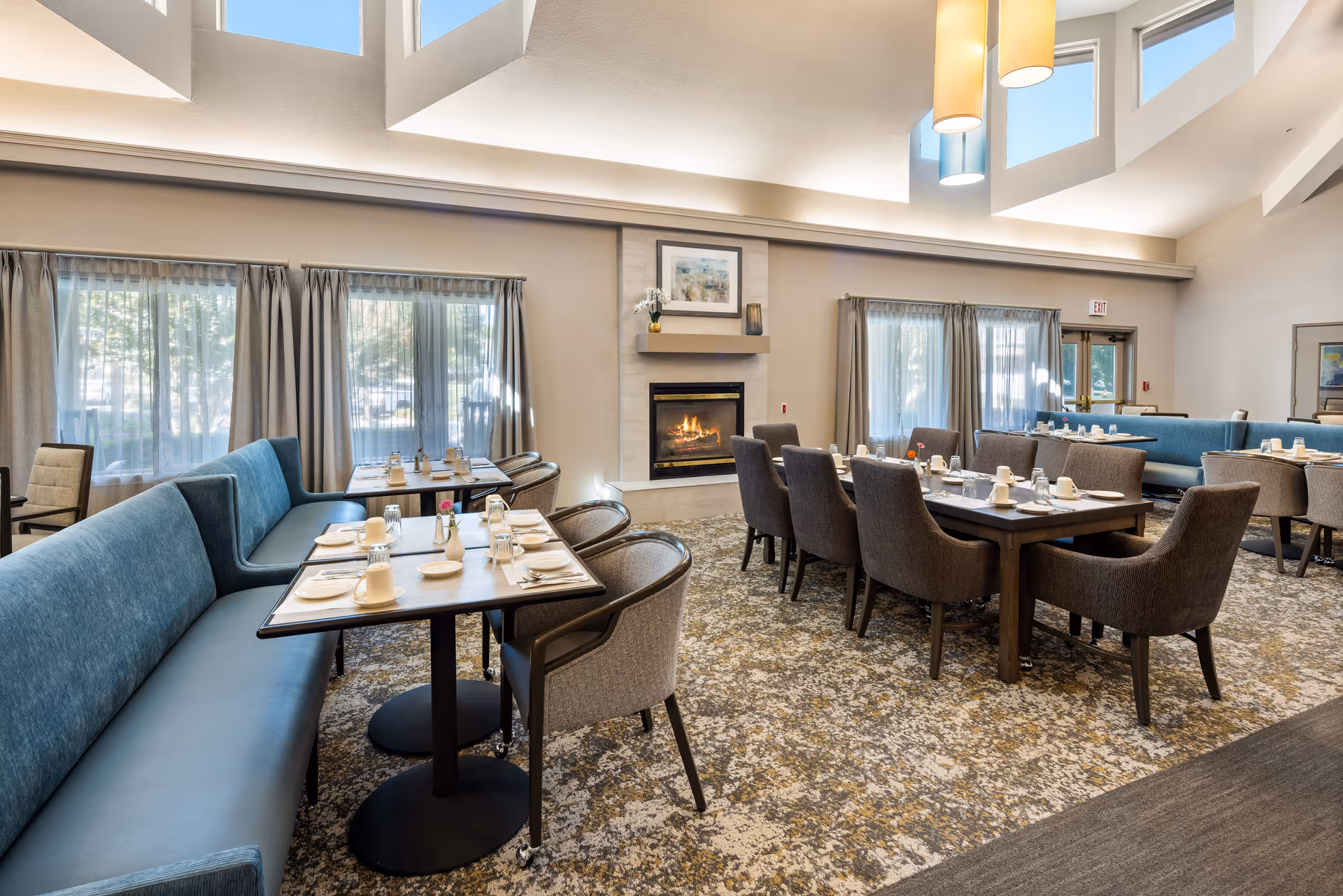 A spacious dining room in a senior living community with multiple tables set for meals. The room features comfortable upholstered chairs and blue cushioned bench seating. Large windows with curtains allow natural light to fill the space. A fireplace with a framed picture above it is centered on one wall, and modern pendant lights hang from the high ceiling.