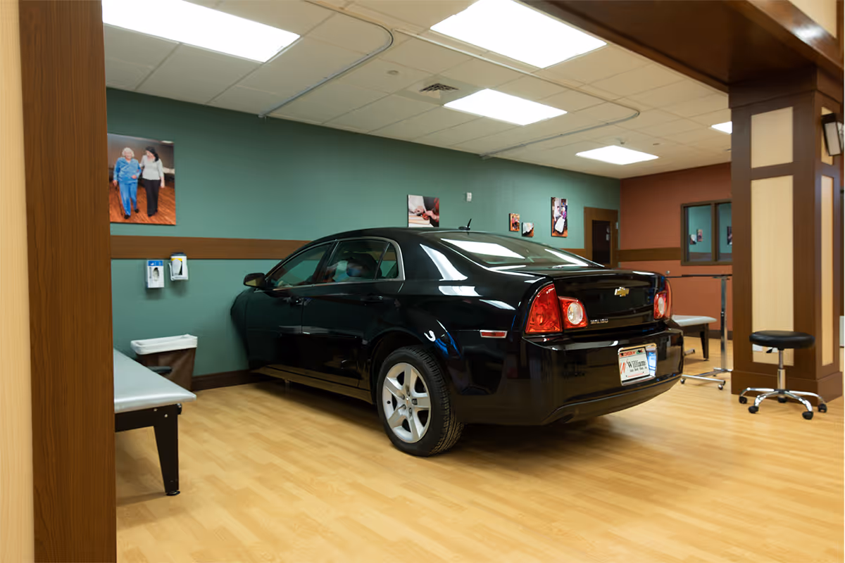 A black sedan parked inside a wood-floored interior room with teal walls, medical/therapy equipment, and framed photos on the walls.