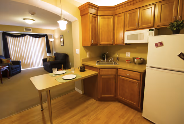 A small kitchenette with wooden cabinets, sink, microwave, refrigerator and a breakfast counter set for two opening into a living area with armchairs and vertical blinds.