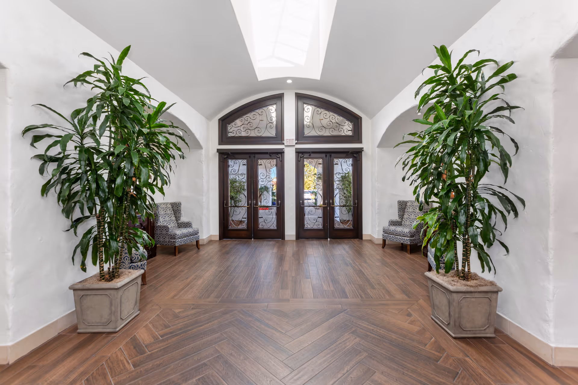 A bright interior lobby with double decorative glass doors, large potted plants, patterned chairs and a skylight above.