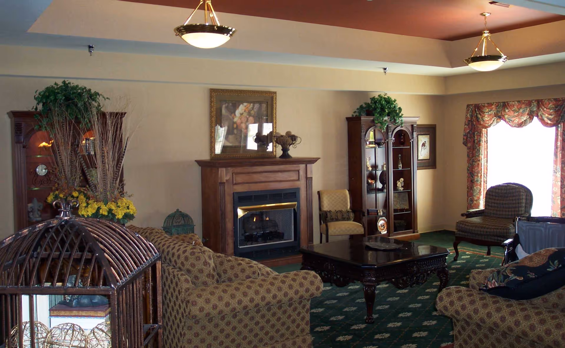 A cozy living room with patterned sofas, a dark wooden coffee table, two armchairs, a fireplace with a framed painting above it, and two wooden display cabinets with decorative items. The room has a green carpet with a floral pattern, beige walls, and a window with floral curtains letting in natural light.