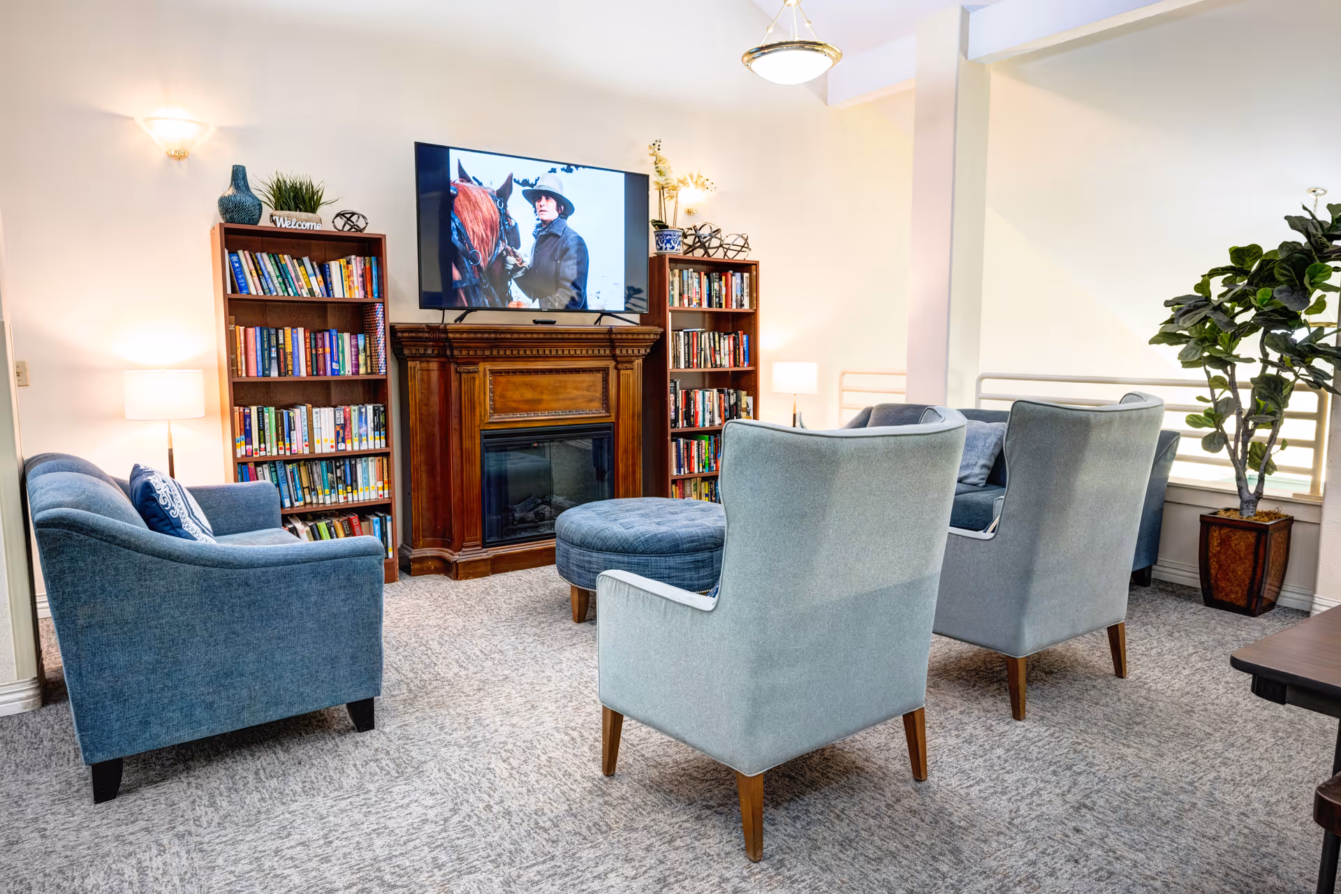 A cozy living room area with blue upholstered chairs and a matching ottoman arranged around a wooden fireplace with a TV mounted above it. Two wooden bookshelves filled with books flank the fireplace. The room is softly lit with wall sconces and table lamps, and there is a large potted plant near a window with white blinds.