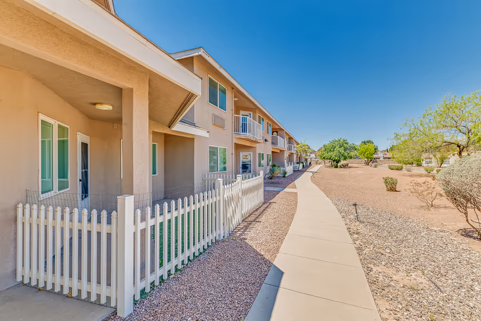 Exterior walkway along a two-story beige apartment building with white picket fences and desert landscaping under a clear blue sky.