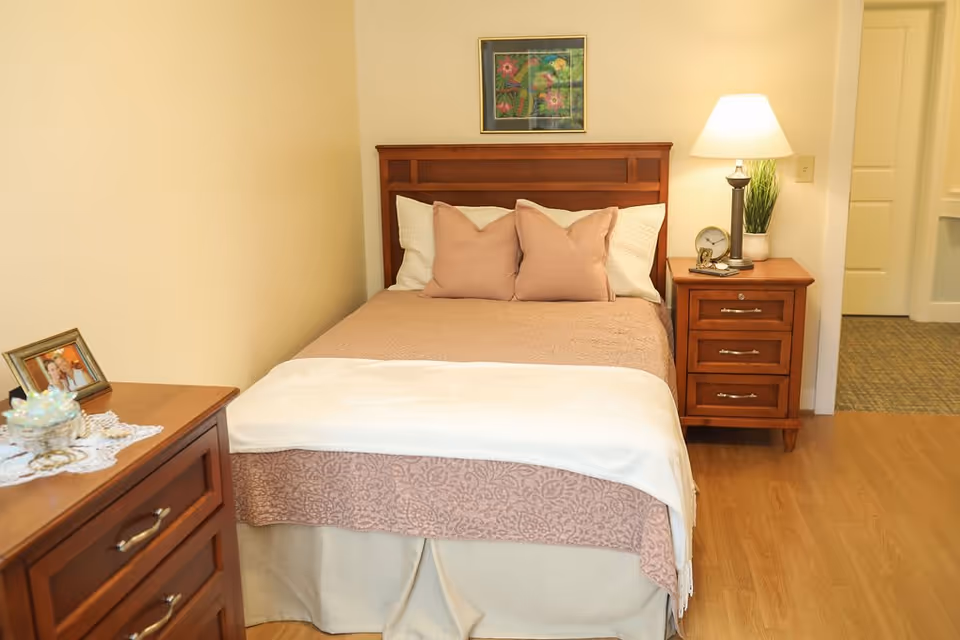 A cozy bedroom with a wooden bed frame, a bed made with beige and white bedding, two beige pillows, a wooden nightstand with a lamp, a small plant, and a clock. There is a wooden dresser with a framed photo and decorative items on it. A colorful framed artwork hangs on the wall above the bed.