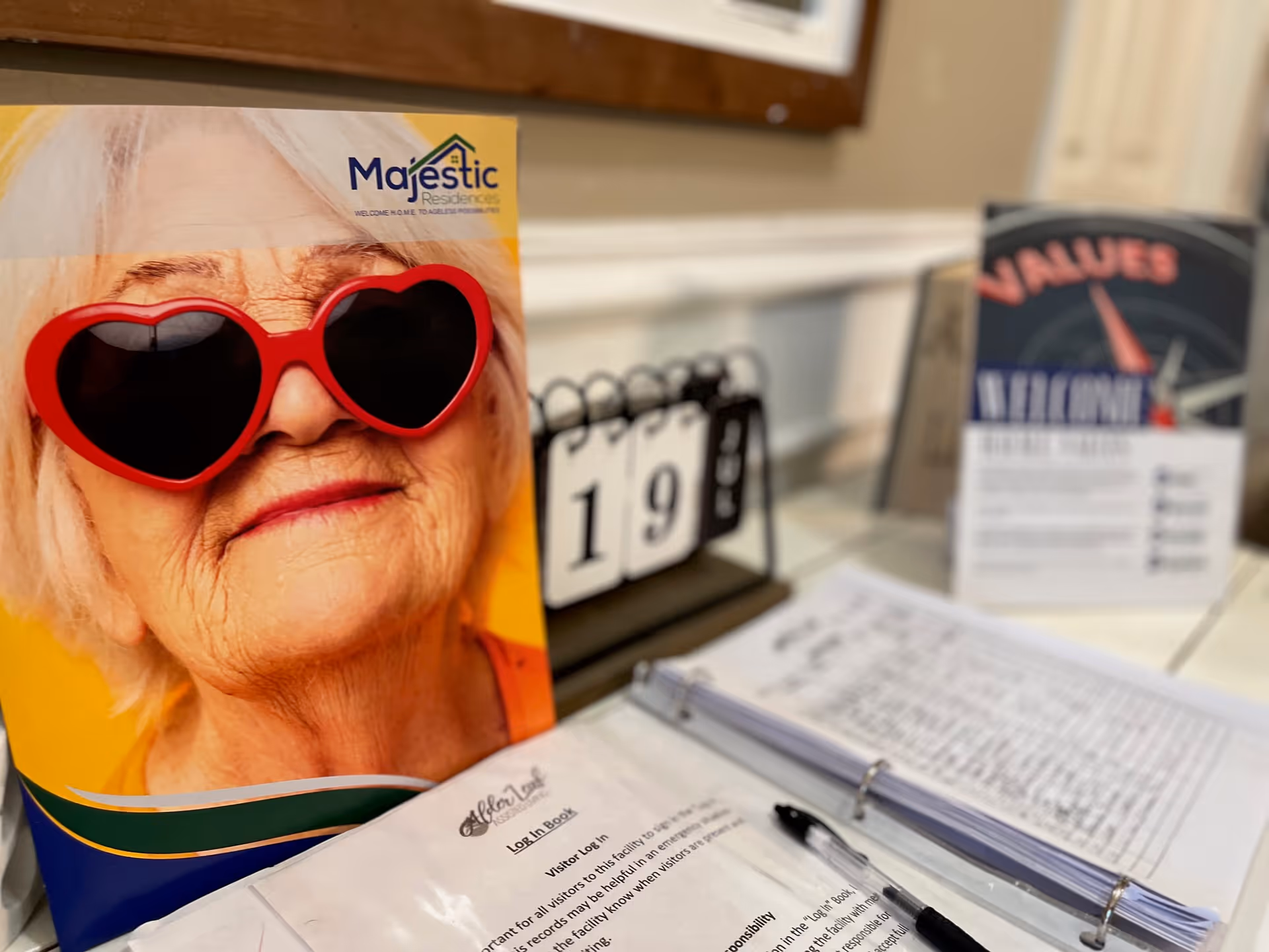 A close-up of a colorful brochure featuring an elderly woman wearing red heart-shaped sunglasses, placed on a counter next to a visitor log book with a pen and a small flip calendar showing the number 19. In the background, there is a blurred sign with the word 'VALUES' visible.