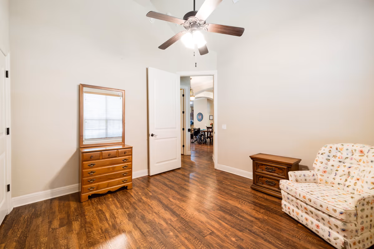 A room with wooden flooring, a ceiling fan with lights, a wooden dresser with a mirror, a small wooden side table, and a floral patterned armchair. The door is open, showing a glimpse of another room with a wheelchair and dining area.