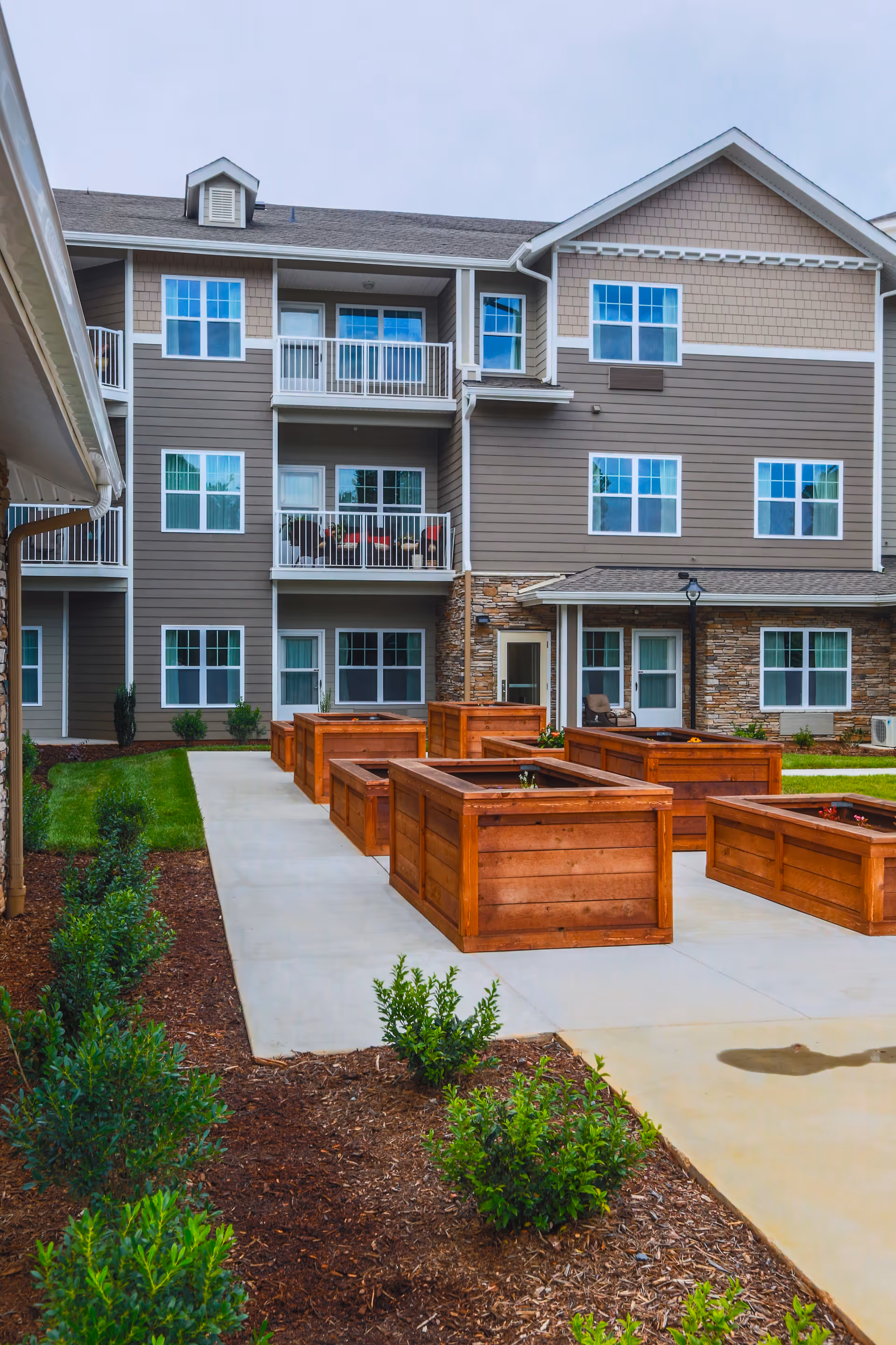 Outdoor courtyard area of a senior living facility with multiple wooden raised garden beds on a concrete patio, surrounded by green shrubs and plants, with a three-story building featuring balconies and windows in the background.