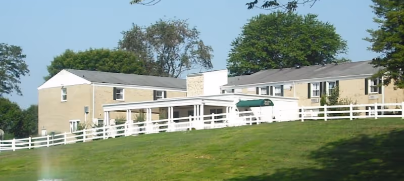 Exterior view of a two-story assisted living facility building with beige brick walls, white trim, and a white fence in front. There is a covered entrance with a green awning and trees in the background under a clear blue sky.