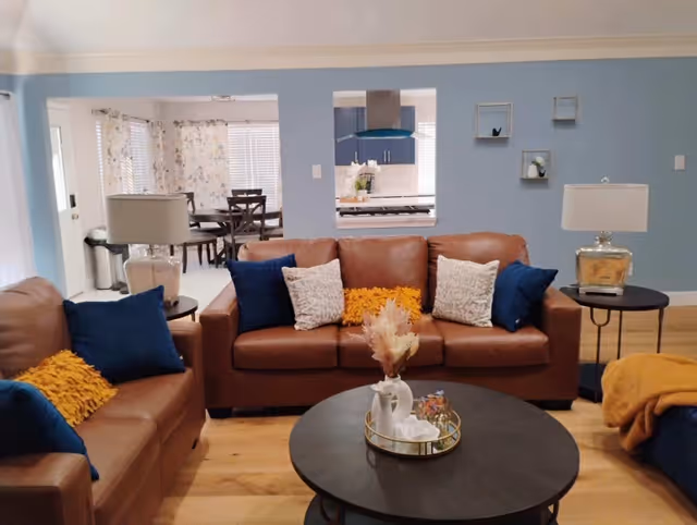 A cozy living room with two brown leather sofas adorned with blue, white, and yellow pillows. A round black coffee table with decorative items sits in the center. The walls are painted light blue with small wall shelves holding plants. Two side tables with lamps flank the sofas. In the background, a dining area with a wooden table and chairs is visible, along with a kitchen area through a pass-through window.