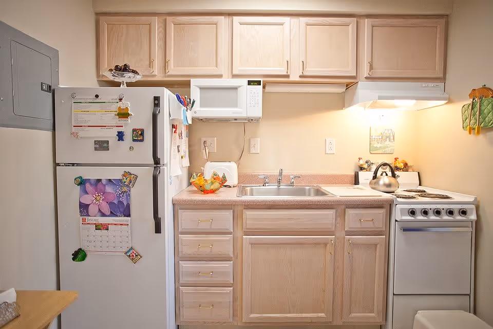 Small kitchen area with light wood cabinets, a white refrigerator with magnets and a calendar, a microwave above the sink, a toaster, a stove with a kettle, and a countertop with a decorative bowl.