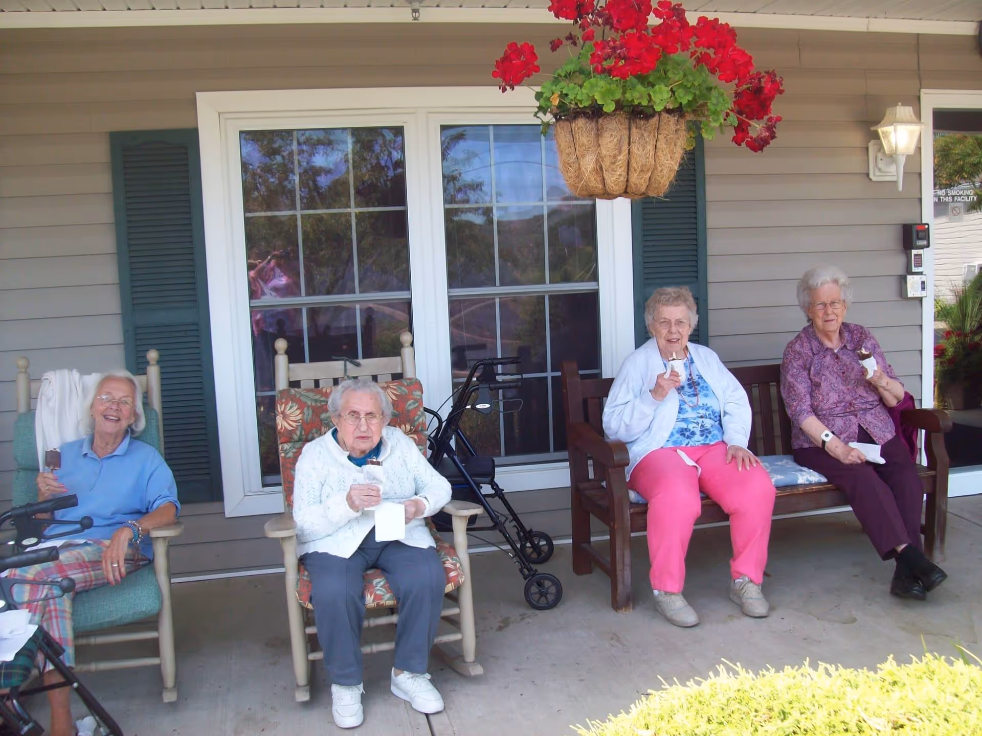 Four elderly women sitting outside on chairs and a bench in front of a building with beige siding and green shutters. They are enjoying ice cream cones under a hanging basket of red flowers.