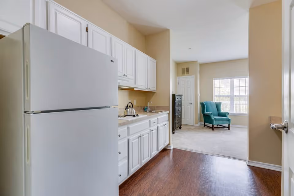 Interior view of a senior living facility kitchen area with white cabinets, a white refrigerator, a countertop with a kettle, and a sink. The kitchen opens into a living room area with a teal armchair near a window with blinds, beige walls, and carpeted flooring.