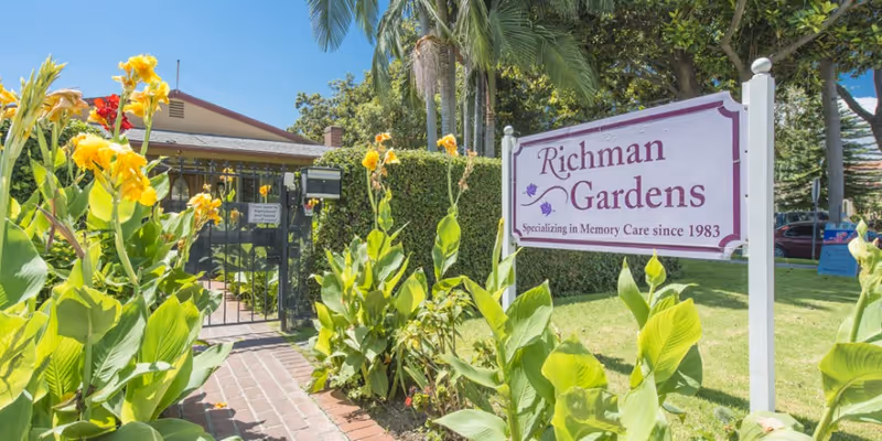Entrance to Richman Gardens facility with a white sign displaying the name and specialization in memory care since 1983, surrounded by green plants and yellow flowers, with a gated walkway and a building partially visible in the background under a clear blue sky.