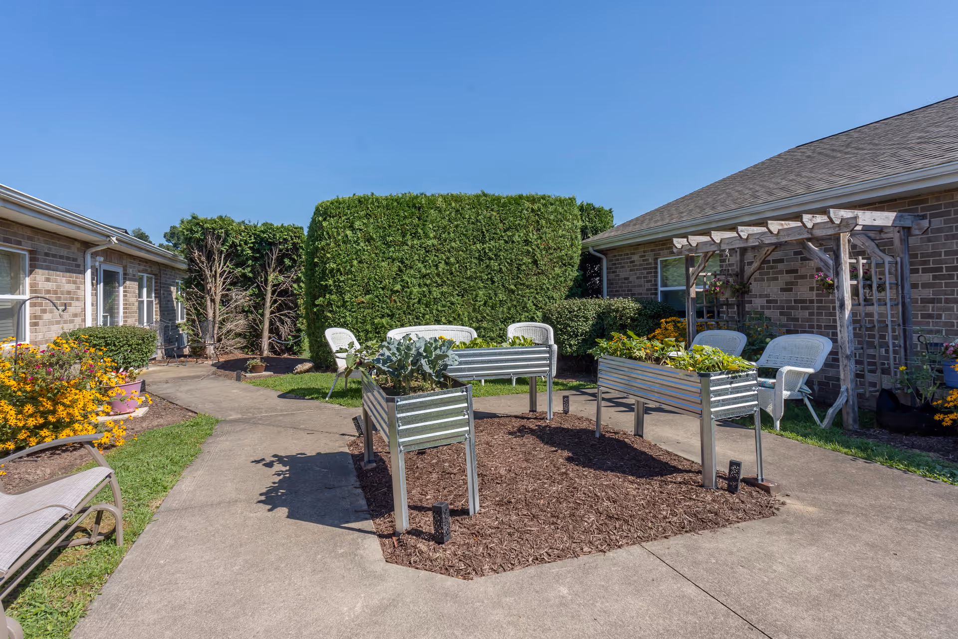 Outdoor garden area with raised metal planters containing various plants, surrounded by a concrete pathway. There are white wicker chairs and a wooden pergola with hanging flower pots near a brick building. The sky is clear and blue.
