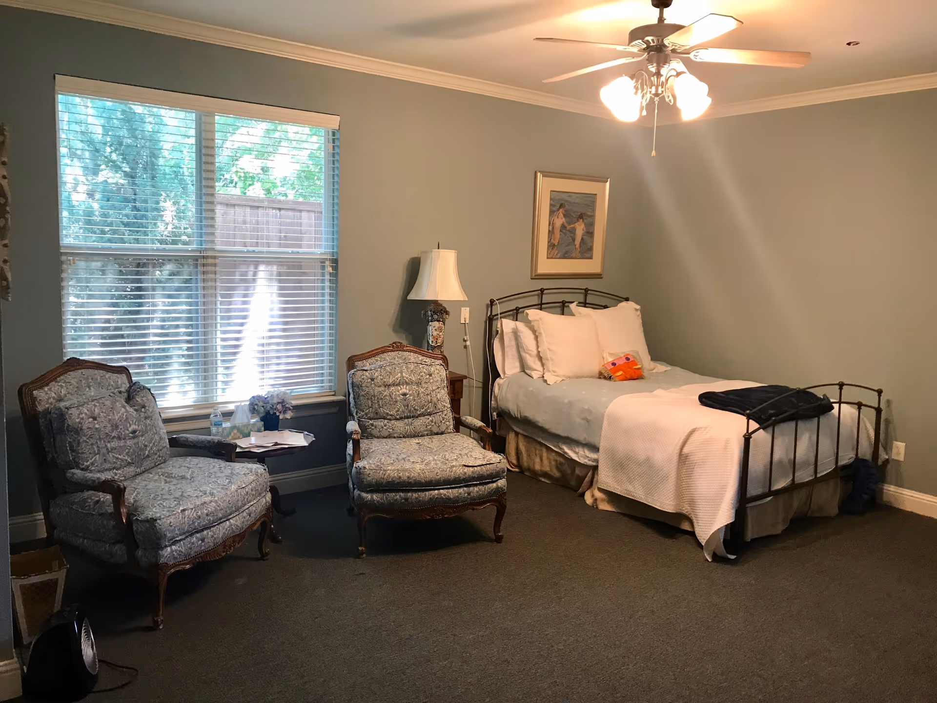 A tranquil bedroom with a metal-framed bed, two upholstered armchairs by a large window, a ceiling fan, and soft blue-green walls.