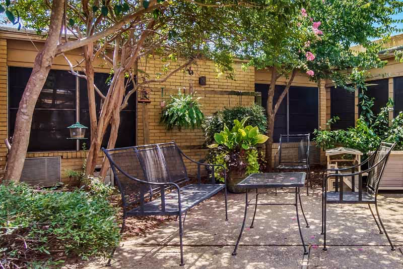 Outdoor patio area with metal chairs and a table surrounded by green plants and trees, adjacent to a brick building with black window coverings.