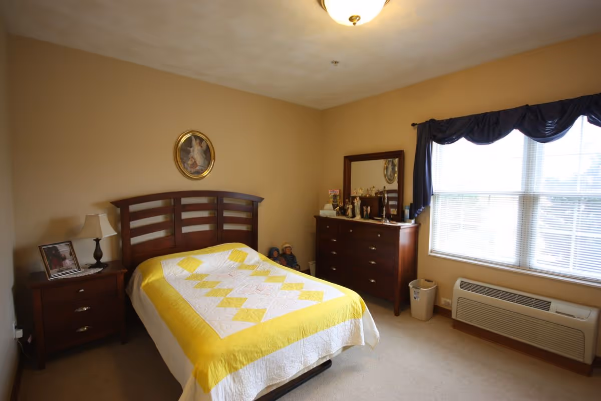 Bedroom with a bed covered by a yellow-and-white quilt, wooden headboard, nightstand with lamp and a dresser beside a large window.