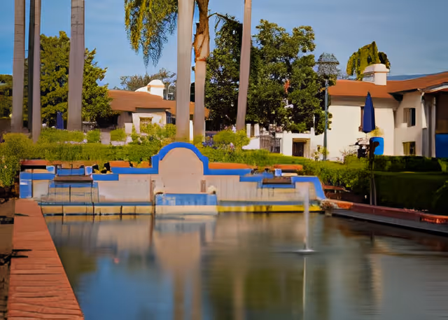 A landscaped courtyard with a rectangular reflecting pool and small fountain in front of low-rise white buildings and tall palm trees.