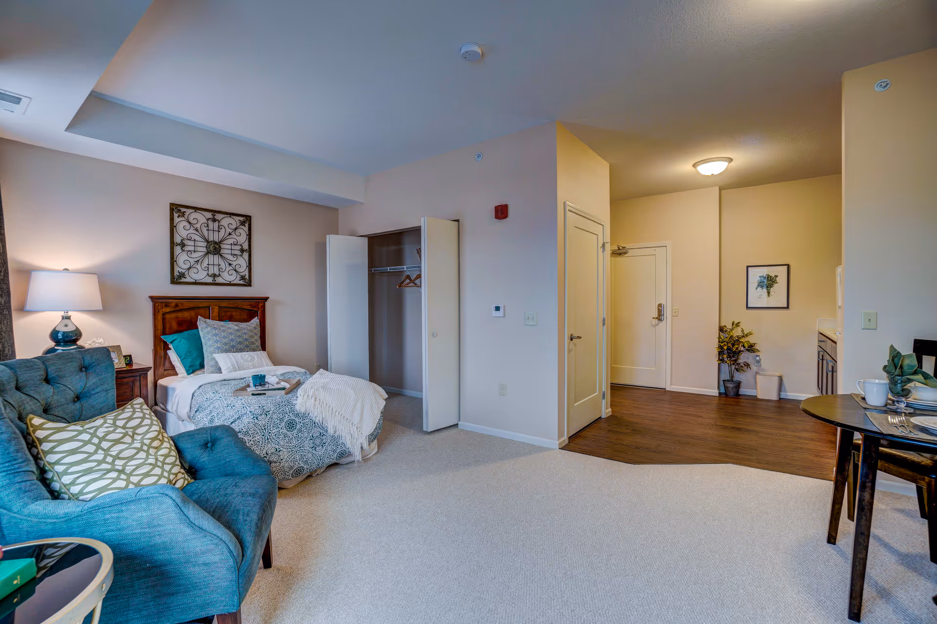 Studio-style apartment interior showing a teal armchair and a single bed beside an open closet, with the entryway and kitchenette visible.