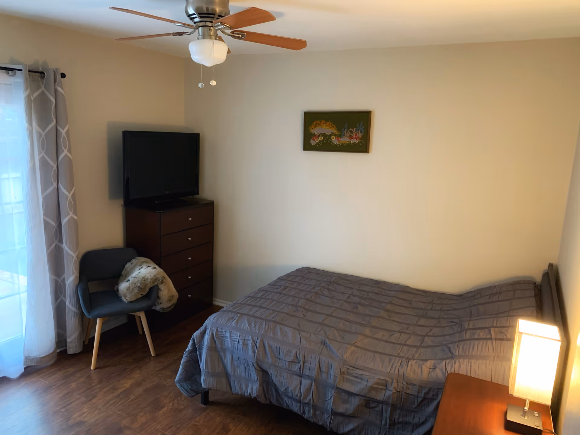 A bedroom with a single bed covered with a gray comforter, a wooden nightstand with a lit lamp, a dark wooden dresser with a flat-screen TV on top, a gray chair with a fur throw, and a ceiling fan with wooden blades. There is a window with sheer and patterned curtains, and a framed floral artwork on the beige wall.