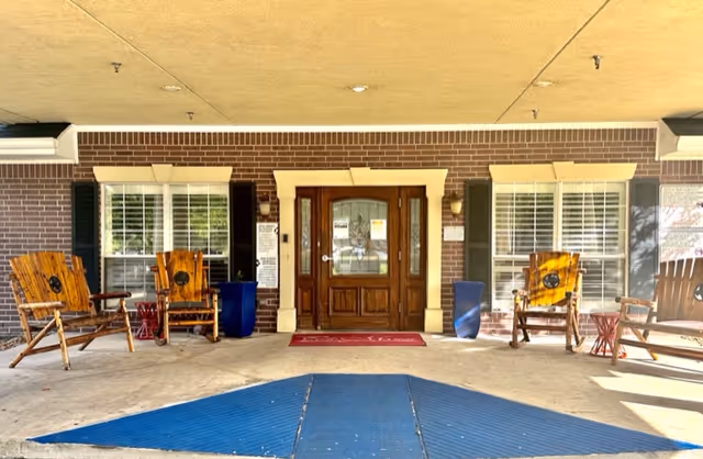 Entrance of a building with a wooden double door framed by brick walls and windows with white shutters on either side. There are wooden rocking chairs and small tables arranged on the covered porch area, with a blue wheelchair ramp leading up to the door.
