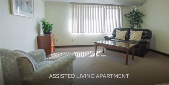 A bright living room in an assisted living apartment with a large window covered by vertical blinds. The room contains a patterned armchair, a black leather loveseat with two decorative pillows, a wooden coffee table with a glass top, and a small wooden cabinet with a potted plant on top. The walls are light-colored and there is a framed picture hanging on the wall.