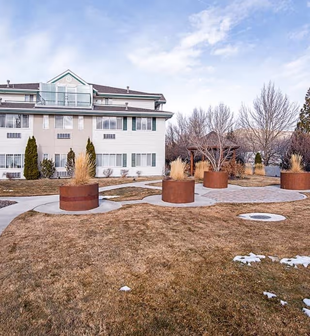 Outdoor view of a senior living facility with a three-story building in the background, a paved walkway, large round planters with tall grasses, leafless trees, and a gazebo. The grass is brown, indicating a winter or early spring season with patches of snow on the ground.