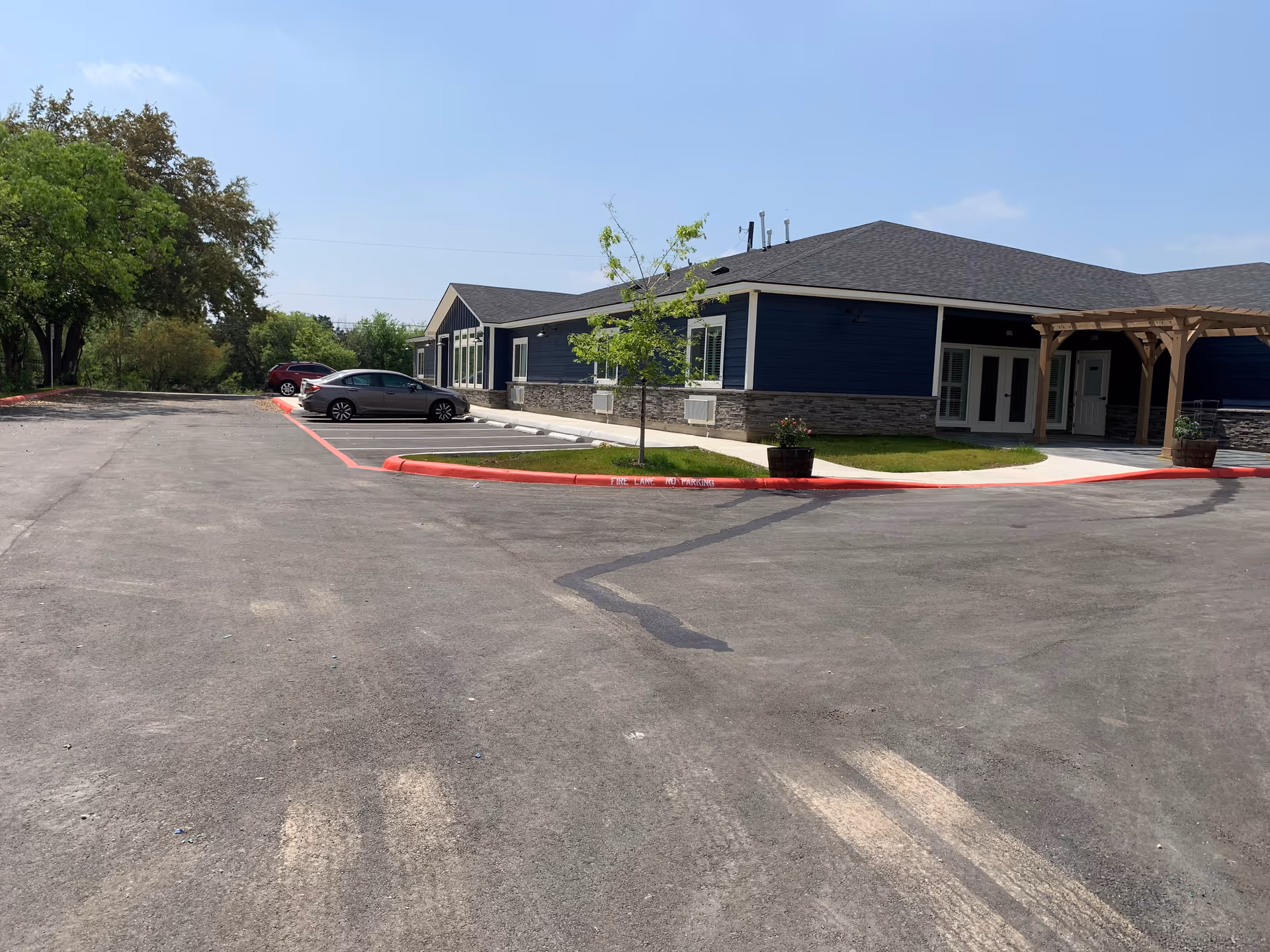 Exterior view of a single-story building with blue siding and stone accents, surrounded by a parking lot with a few parked cars. There are small trees and planters near the building, and a wooden pergola over the entrance. The sky is clear and blue.