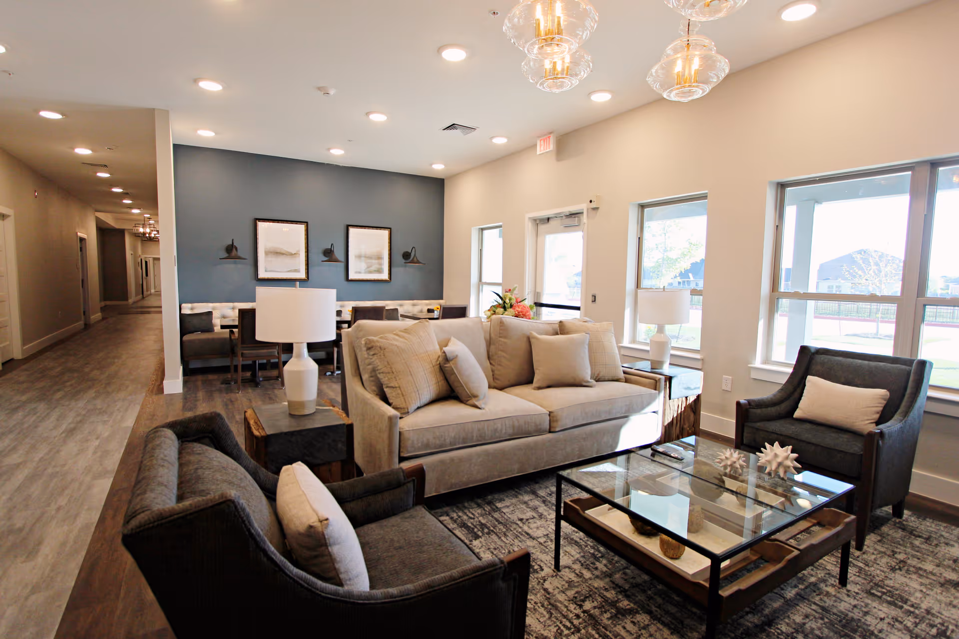 A bright and modern living room area in a senior living facility featuring a beige sofa with multiple cushions, two dark armchairs with light pillows, a glass coffee table with decorative items, and two side tables with white lamps. The room has large windows letting in natural light, a blue accent wall with framed artwork and wall sconces, and a hallway leading to other rooms.