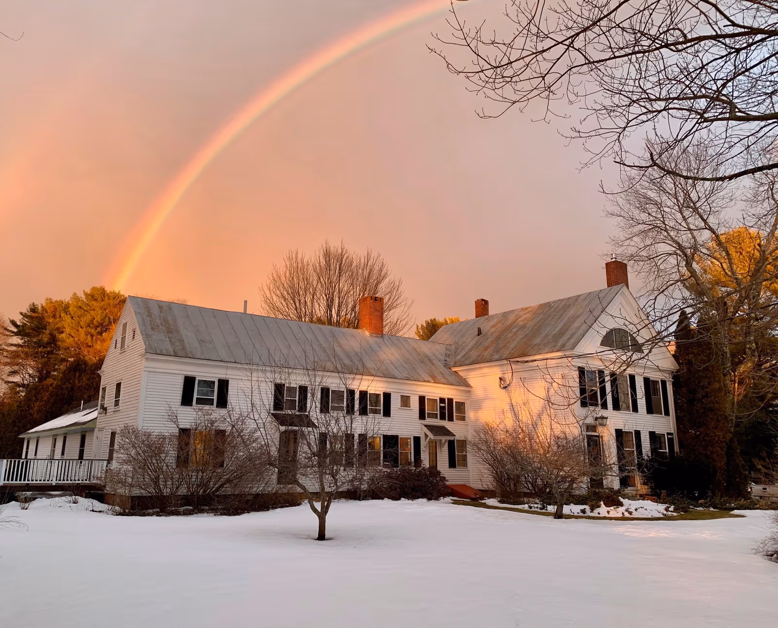 A large white two-story building with black shutters and a metal roof, surrounded by leafless trees and snow-covered ground. A rainbow arcs across the sky above the building during sunset.