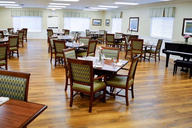 A spacious dining room with multiple wooden tables and chairs arranged neatly on a polished wooden floor. Each table is set with glasses, napkins, and small flower vases. The room has large windows with blinds, framed artwork on the walls, and a black piano in the corner.
