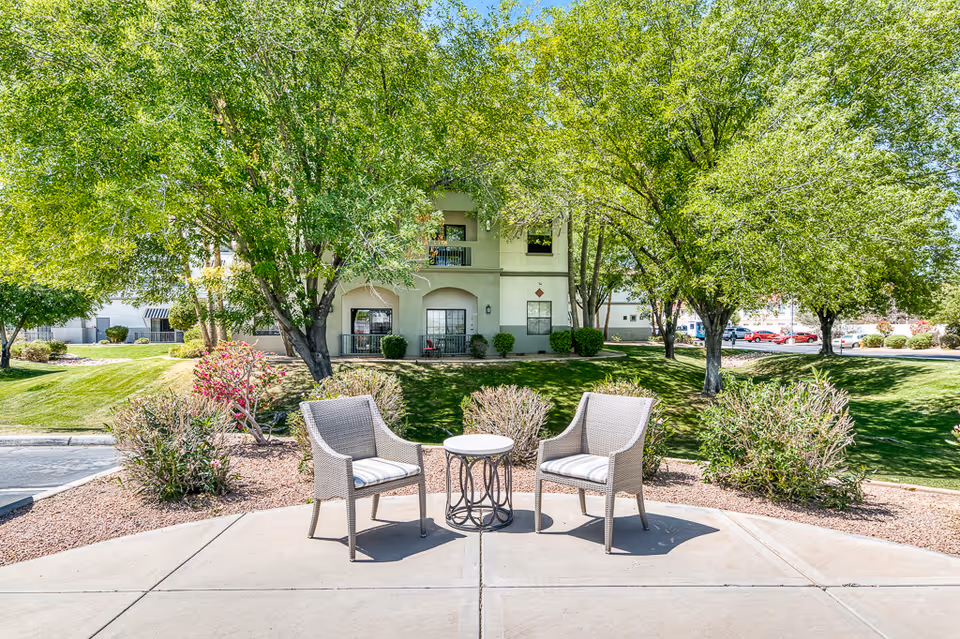 Two wicker chairs with cushions and a small round table between them are placed on a concrete patio area surrounded by landscaping with bushes and trees. In the background, there is a two-story building with balconies and windows, and a well-maintained grassy area with more trees and parked cars visible in the distance under a clear blue sky.