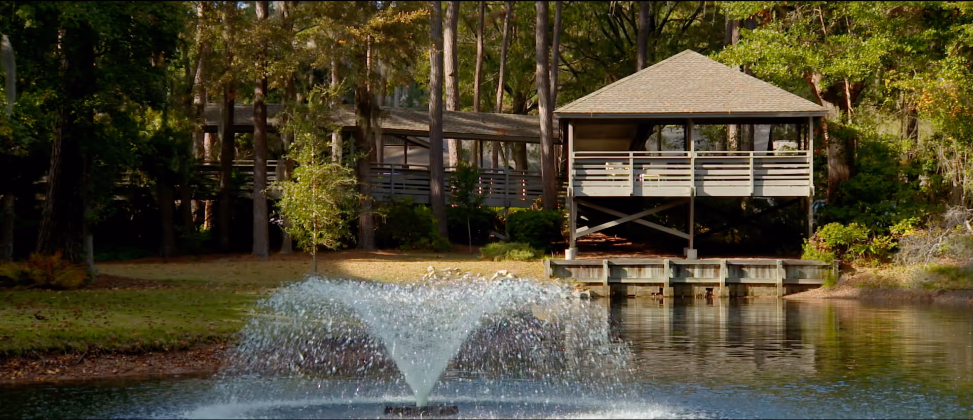 A peaceful outdoor scene featuring a water fountain spraying water in a pond, with a wooden gazebo and a covered walkway surrounded by tall trees and greenery in the background.