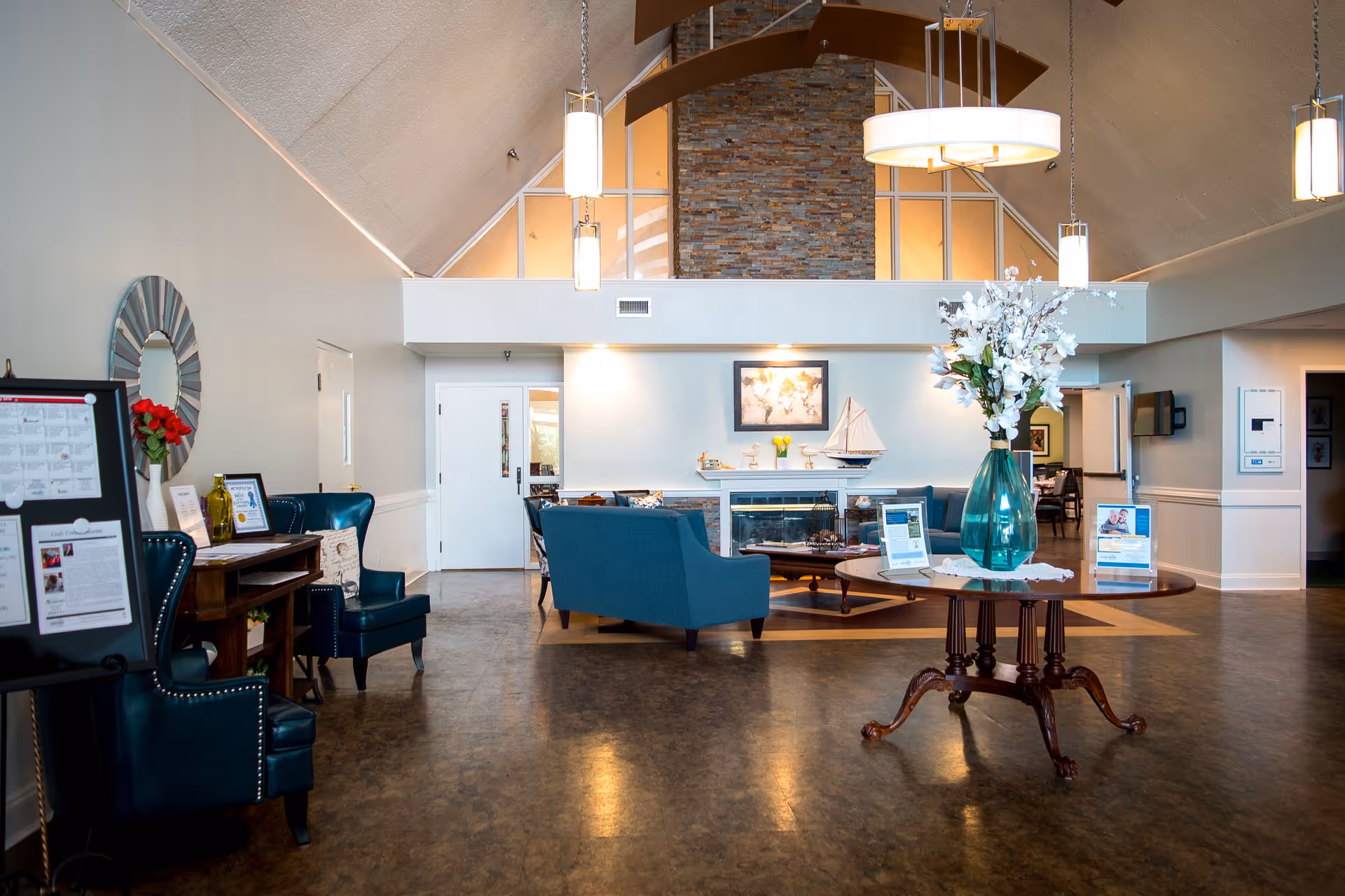 A spacious and well-lit living room area with high vaulted ceilings and a stone fireplace. The room features blue upholstered chairs and sofas arranged around the fireplace. A round wooden table with a large vase of white flowers is in the foreground. There are pendant lights hanging from the ceiling and decorative items including a model sailboat on the fireplace mantel. The walls are painted light gray, and there is a mirror and framed pictures on the walls.