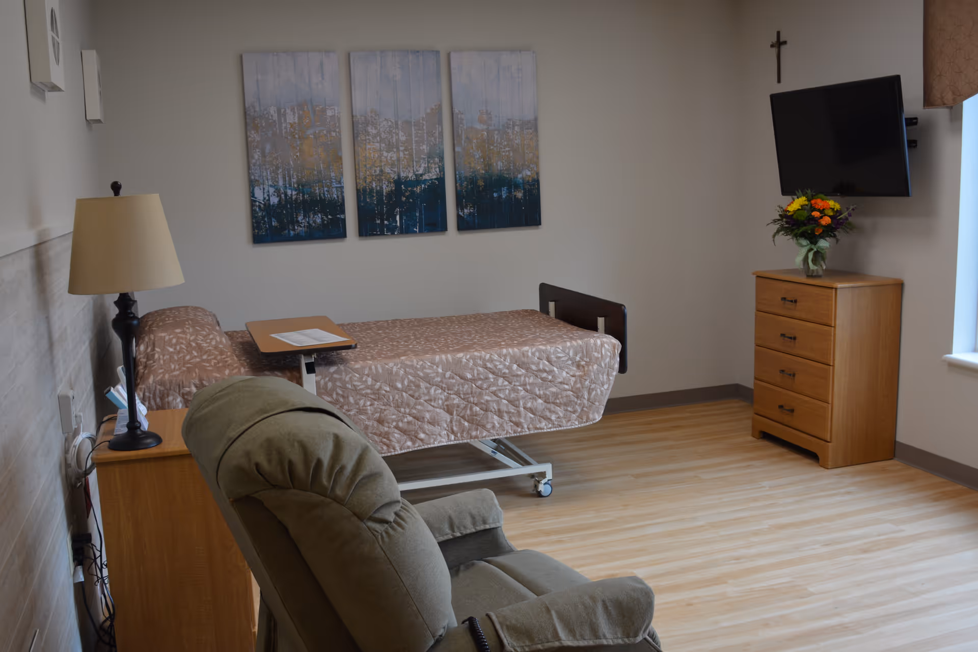 A senior living facility room with a hospital-style bed covered in a patterned quilt, a wooden nightstand with a lamp, a green recliner chair, a wooden dresser with a vase of flowers on top, a wall-mounted flat-screen TV, and a crucifix on the wall. Three abstract paintings hang above the bed, and there is a window with a curtain on the right side.