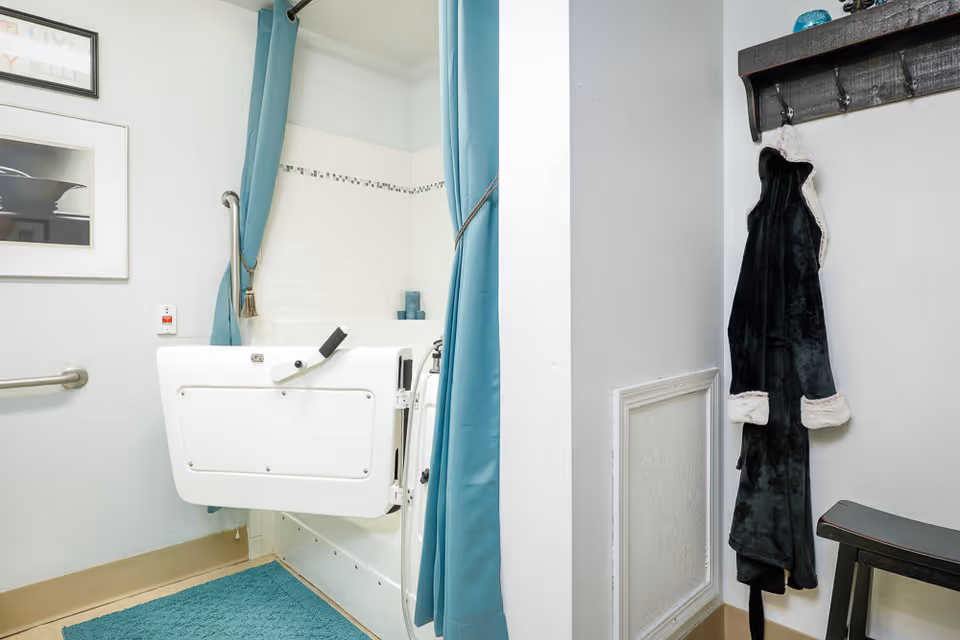 A bathroom with a walk-in bathtub featuring a white door and blue curtains tied back. There is a blue bath mat on the floor, a grab bar on the wall, and a black robe hanging on a hook next to a small bench.