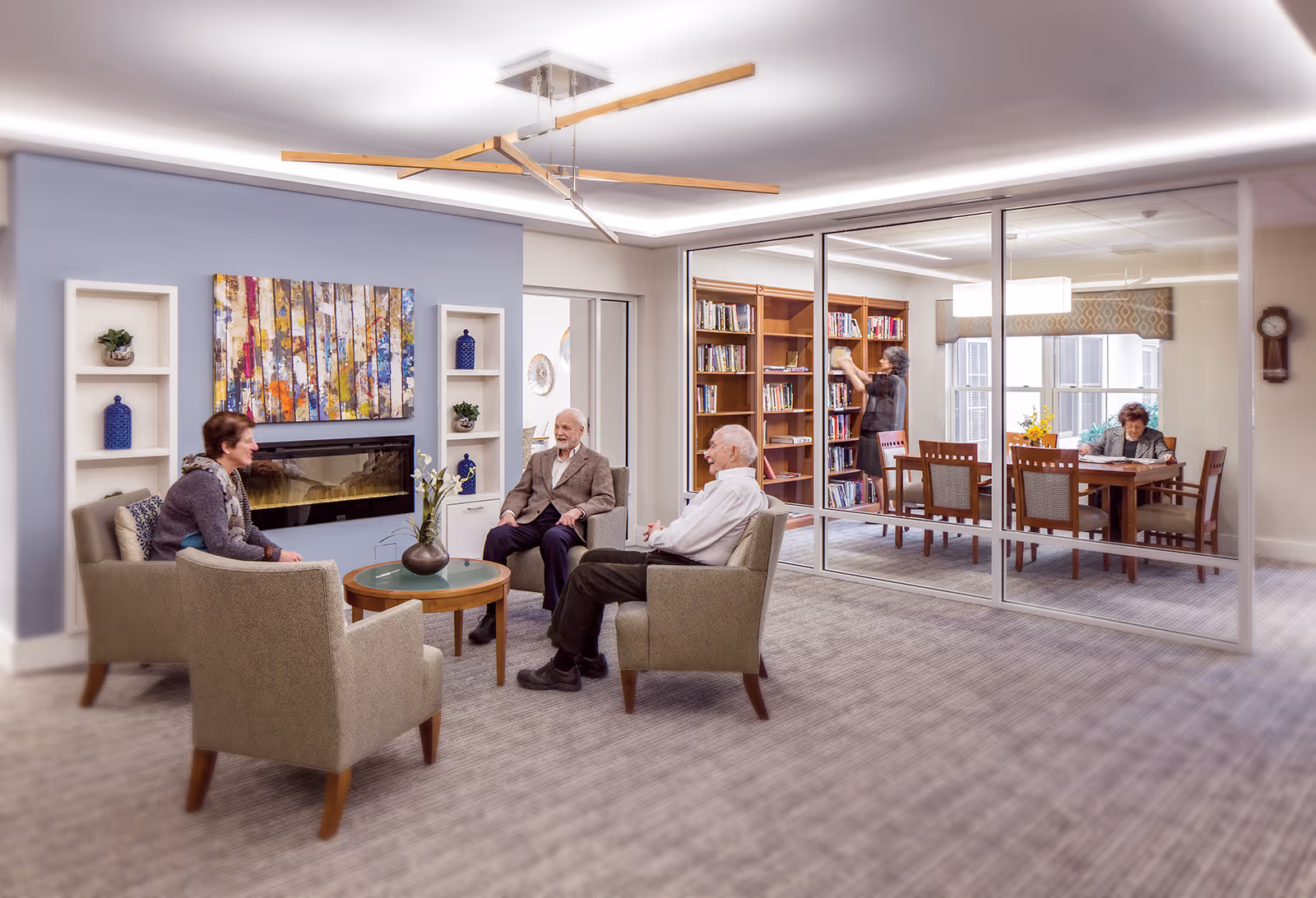 A senior living facility lounge with four armchairs around a coffee table, a wall-mounted fireplace and a glass-walled library/dining room where residents read and browse books.