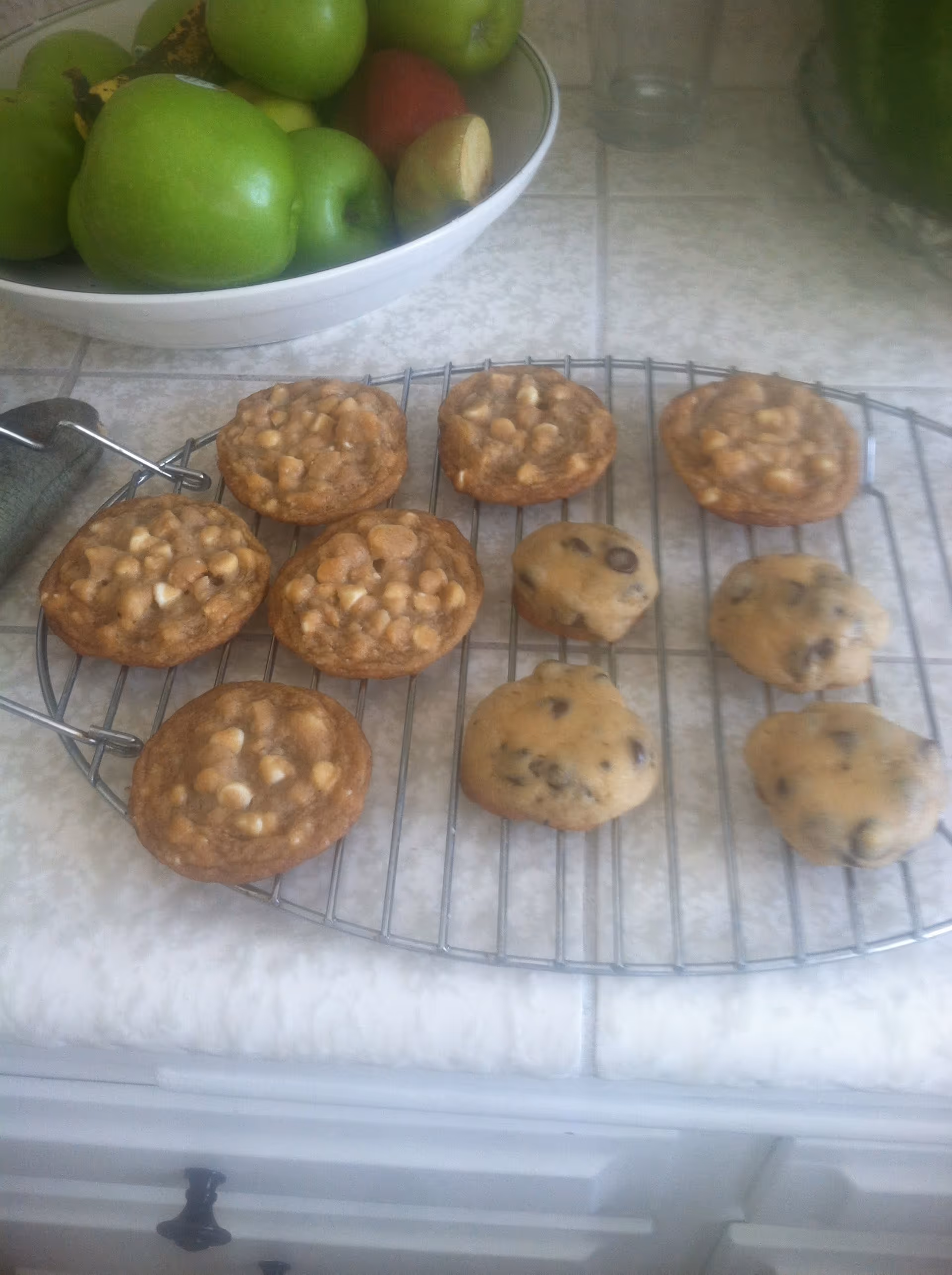 A cooling rack on a kitchen counter with freshly baked cookies, some with white chocolate chips and others with chocolate chips. In the background, there is a white bowl filled with green apples and a few other fruits.