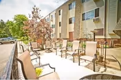 Outdoor patio area with multiple chairs arranged around small tables next to a multi-story residential building with windows. Trees and parked cars are visible in the background under a partly cloudy sky.