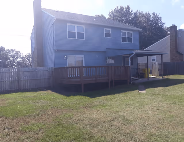 Back view of a two-story blue house with a wooden deck and a covered patio area, surrounded by a fenced grassy yard.