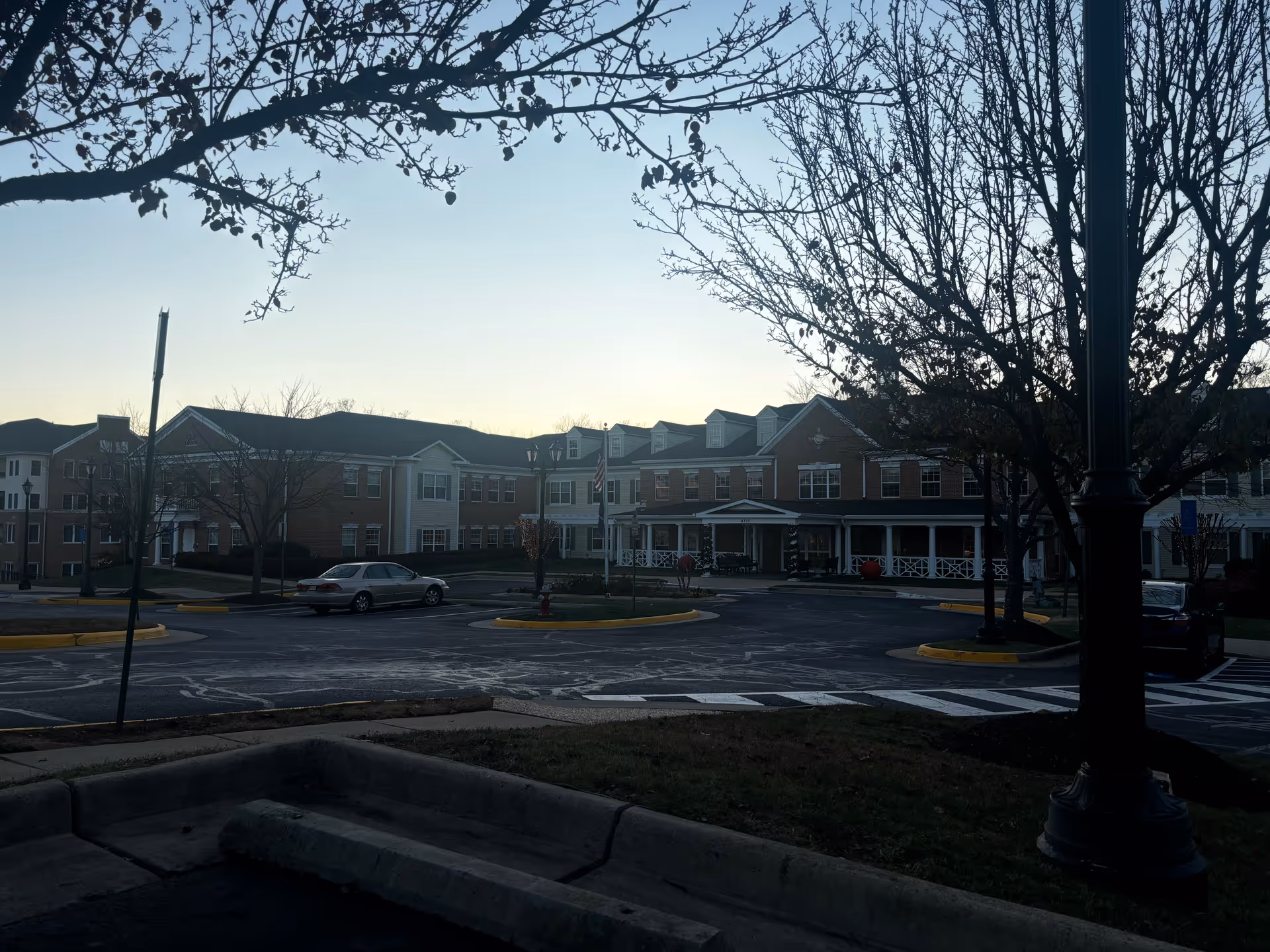 Exterior view of a senior living facility building with a parking lot in front, surrounded by leafless trees and a street lamp during early evening or dawn.
