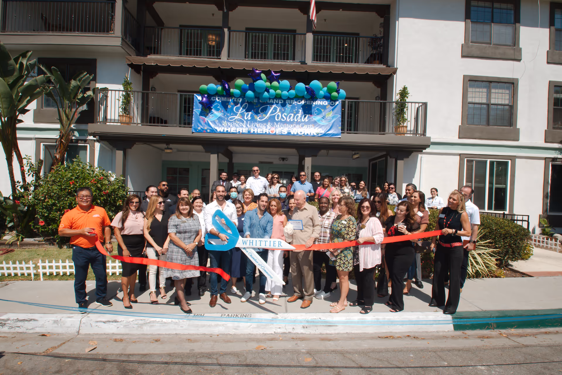 A large group of people gathers in front of La Posada assisted living building for a ribbon-cutting ceremony, holding a red ribbon and oversized scissors beneath a banner.