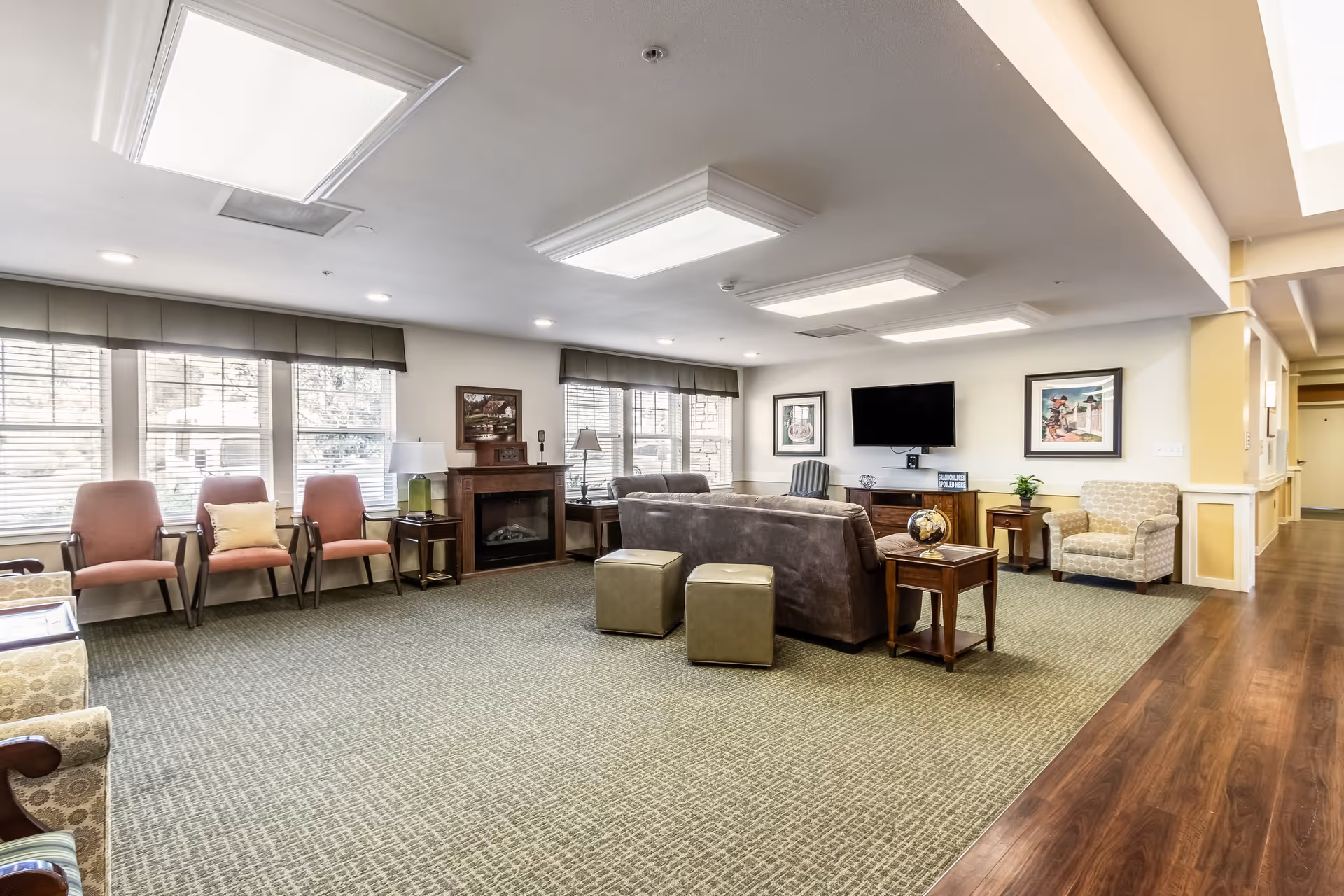 A spacious living room area in a senior living facility with multiple chairs arranged along the walls and around a central brown sofa. The room features large windows with blinds, a fireplace, a wall-mounted flat-screen TV, framed artwork, and side tables with lamps and decorative items. The floor is carpeted with a patterned design, and there is wood flooring along the hallway.