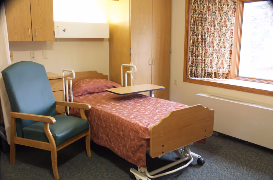A nursing facility room with a single hospital bed covered in a patterned reddish-brown blanket, an overbed table, a green cushioned armchair with wooden arms, wooden cabinets, a window with floral curtains, and a heating unit below the window.
