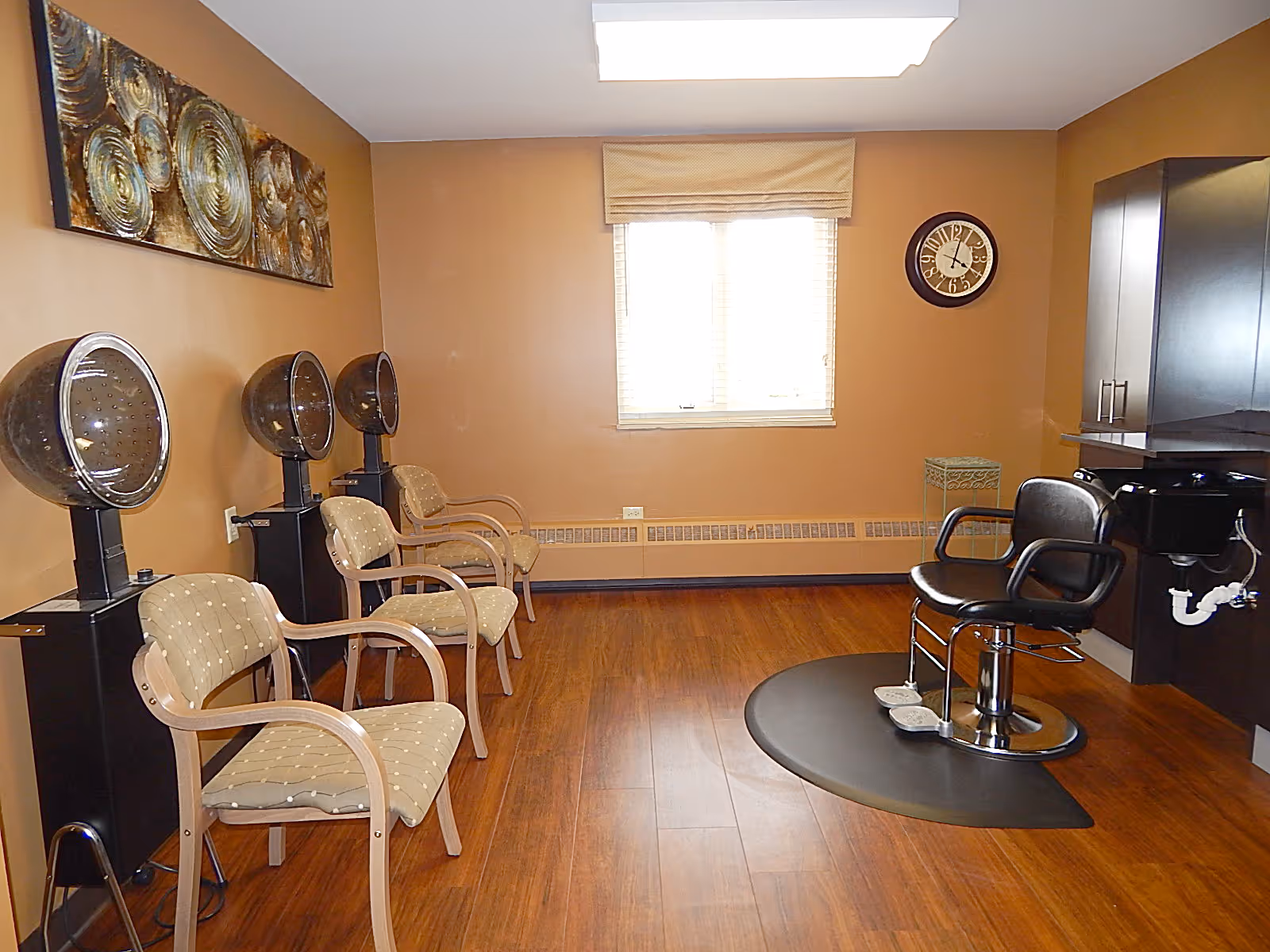 Interior view of a salon area in a senior living facility with three beige cushioned chairs lined up against the left wall, each positioned in front of a hair dryer hood. On the right side, there is a black salon chair on a circular mat in front of a black sink and cabinetry. The room has wood flooring, tan walls, a window with blinds and a valance, a wall clock, and a decorative wall art piece.