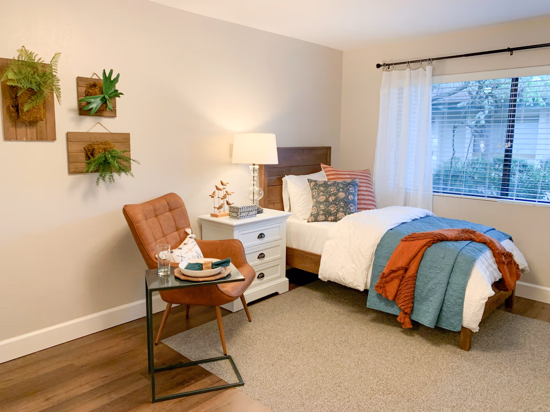 Cozy single bedroom with a bed dressed in teal and orange throws, a nightstand with a lamp, an accent chair, and a window with blinds.