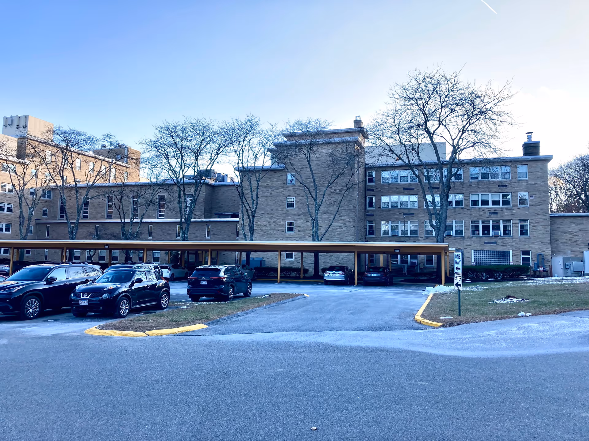 Exterior view of a multi-story brick building with several windows, a covered parking area with cars parked underneath, leafless trees around the building, and a one-way traffic sign near the parking lot entrance.