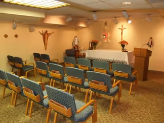 Small chapel room with rows of blue cushioned chairs facing an altar adorned with a white cloth, flowers, and religious statues and crosses on the walls.