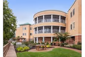 Three-story brick senior living building with rounded balconies overlooking a landscaped courtyard with lawn, seating, and walkways.